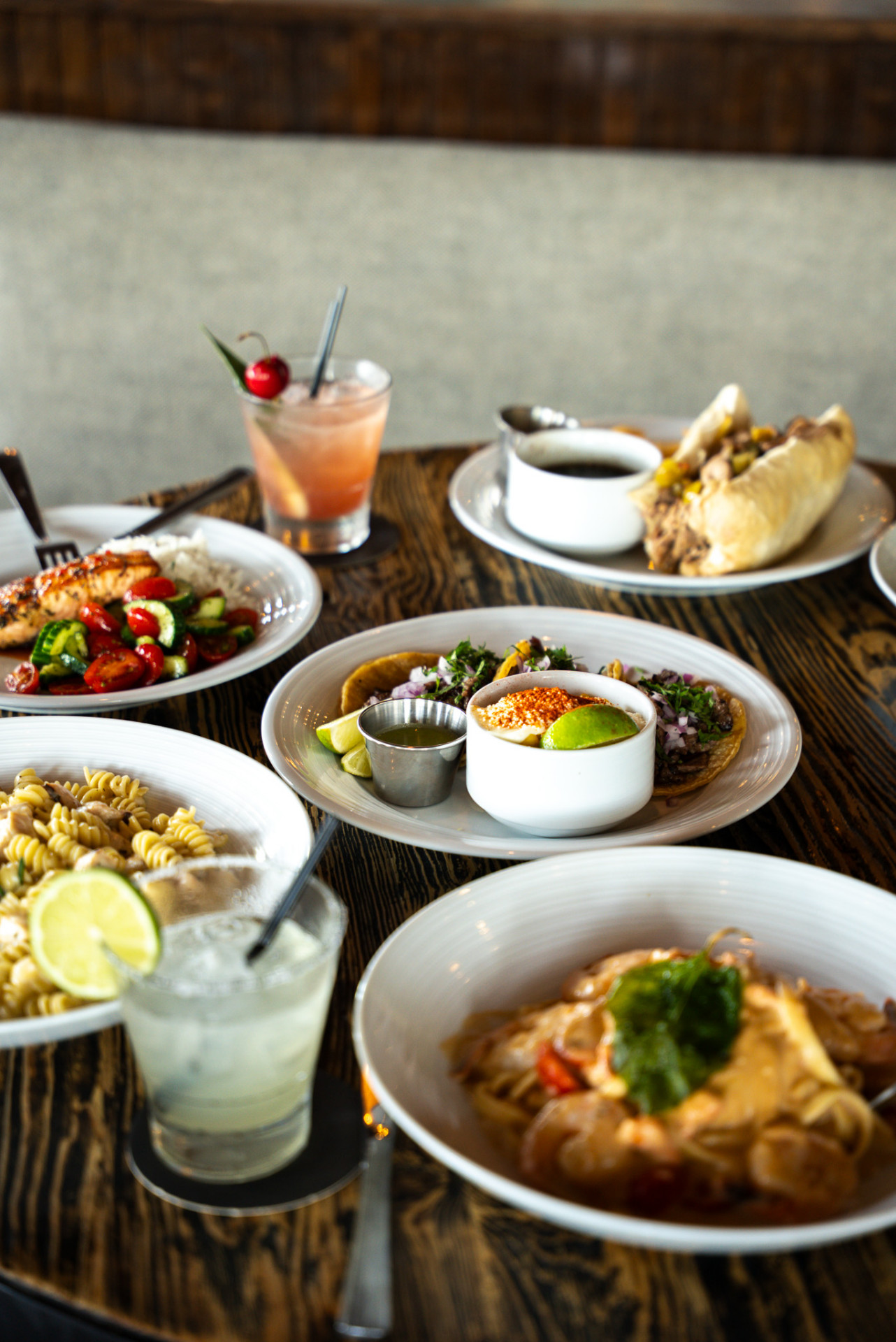 A spread of Urban Kitchen dishes including birria tacos, French dip, salmon, pasta, and cocktails on a dark wood table