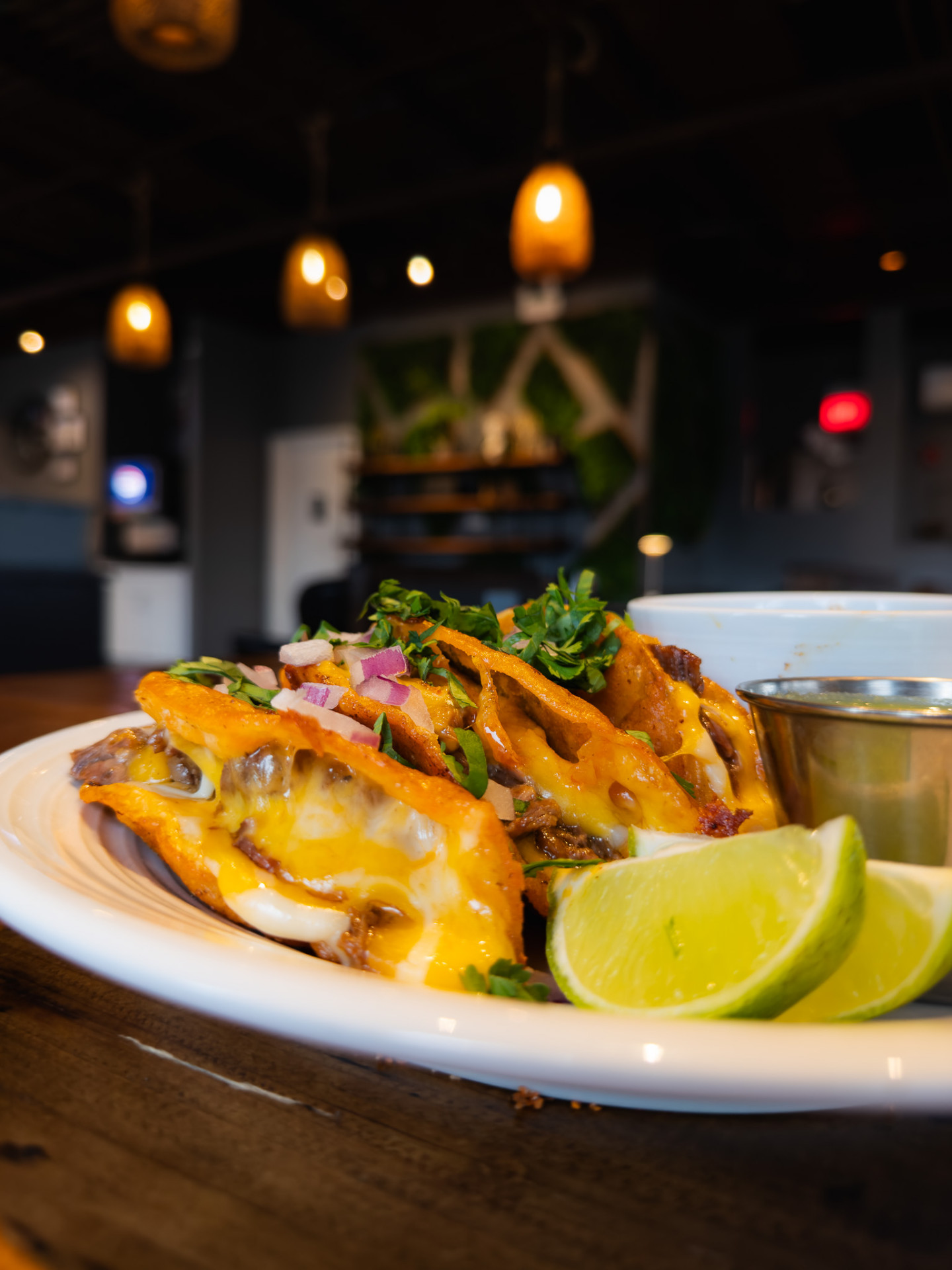 Three crispy birria tacos topped with onion and cilantro, with a side of consommé and lime